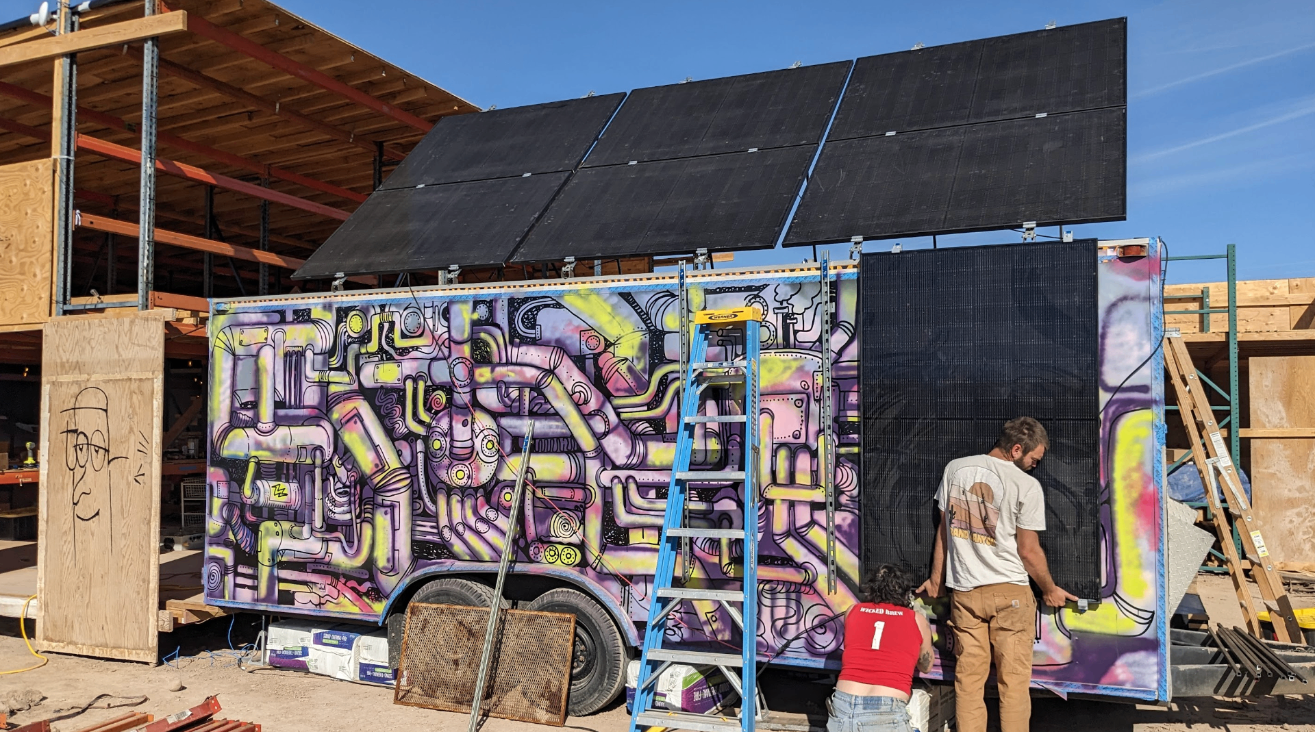 A large mural of intertwined pipes and machinery painted in pink, yellow, and purple on the side of a trailer in the desert