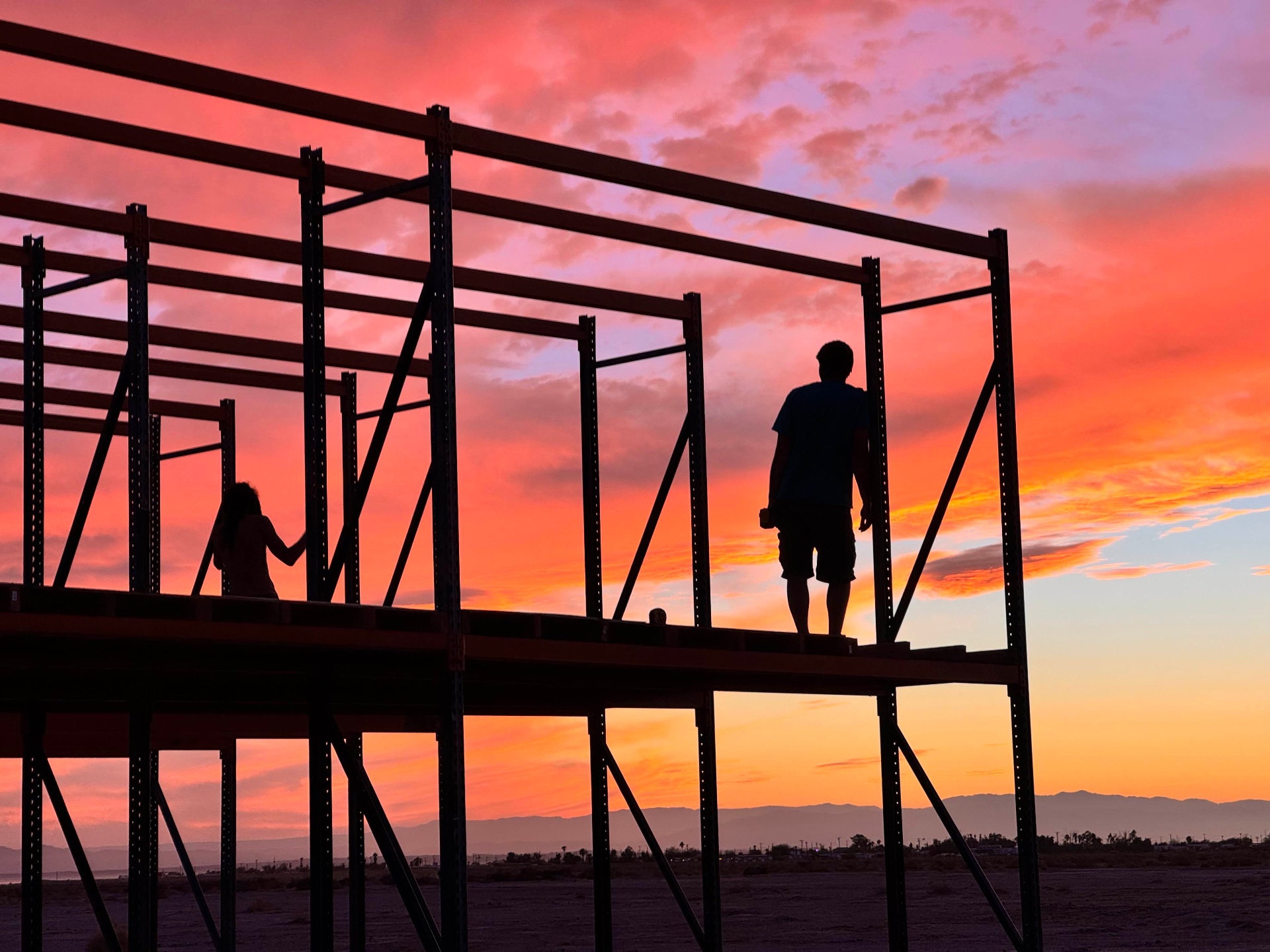Two people silhouetted on a pallet-rack structure against a vivid pink and orange desert sunset