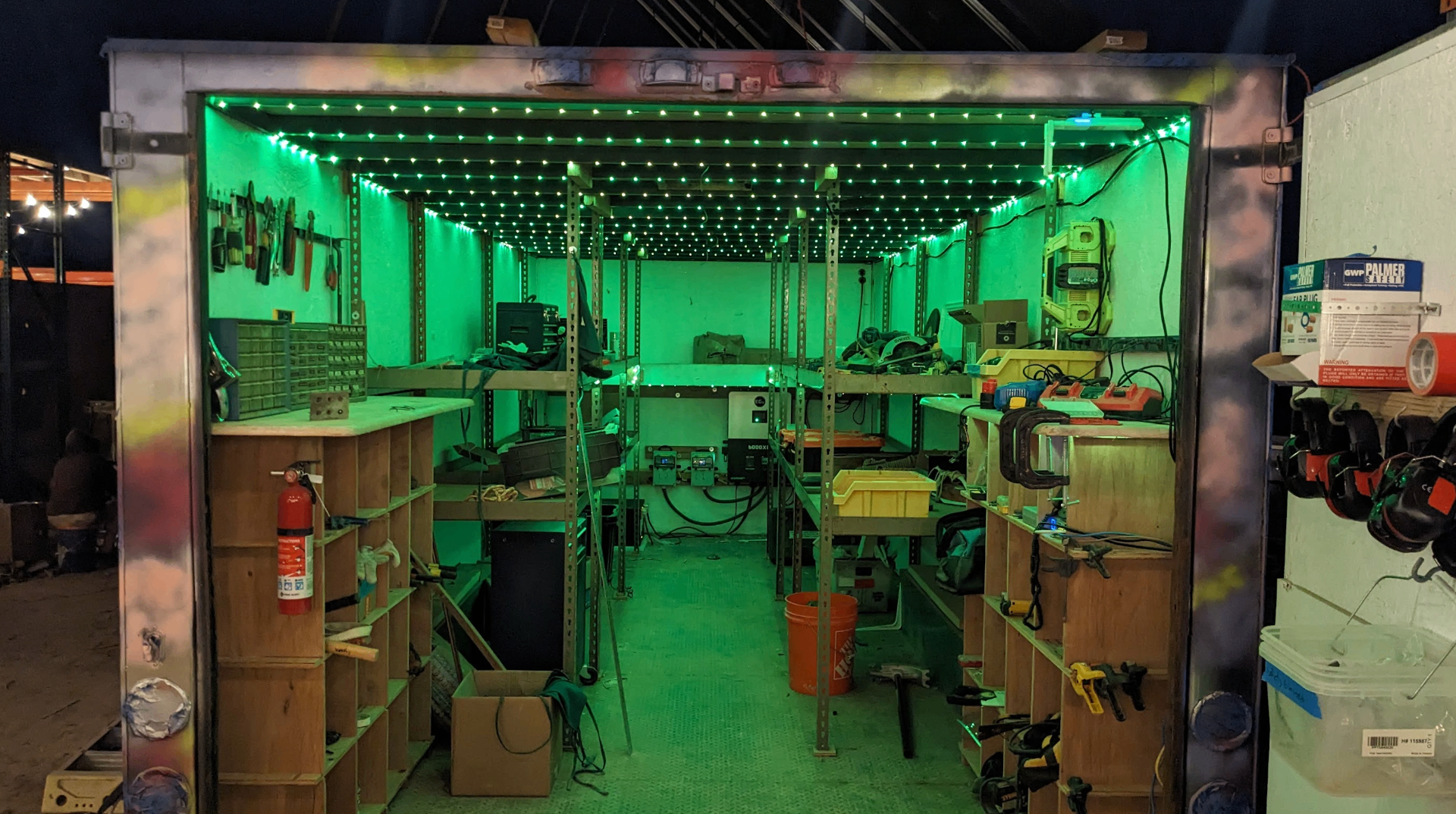 Inside the workshop trailer at night, glowing green LED lights above shelves of tools and equipment